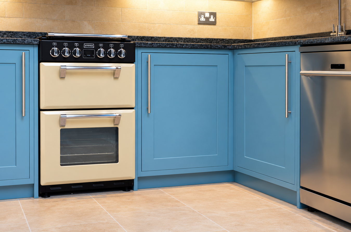 A kitchen corner with bright blue shaker-style cabinets, a cream double oven stove, dark speckled countertops, beige tiled walls and floor, and part of a stainless steel dishwasher visible on the right.